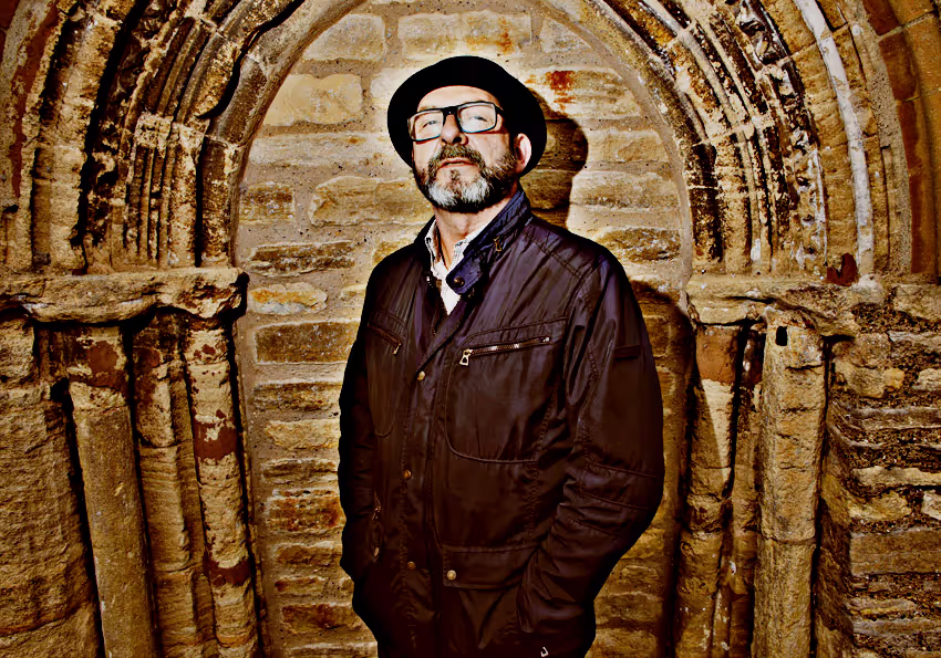 A man wearing a black jacket and glasses stands confidently in front of a stone archway, showcasing a historic backdrop.