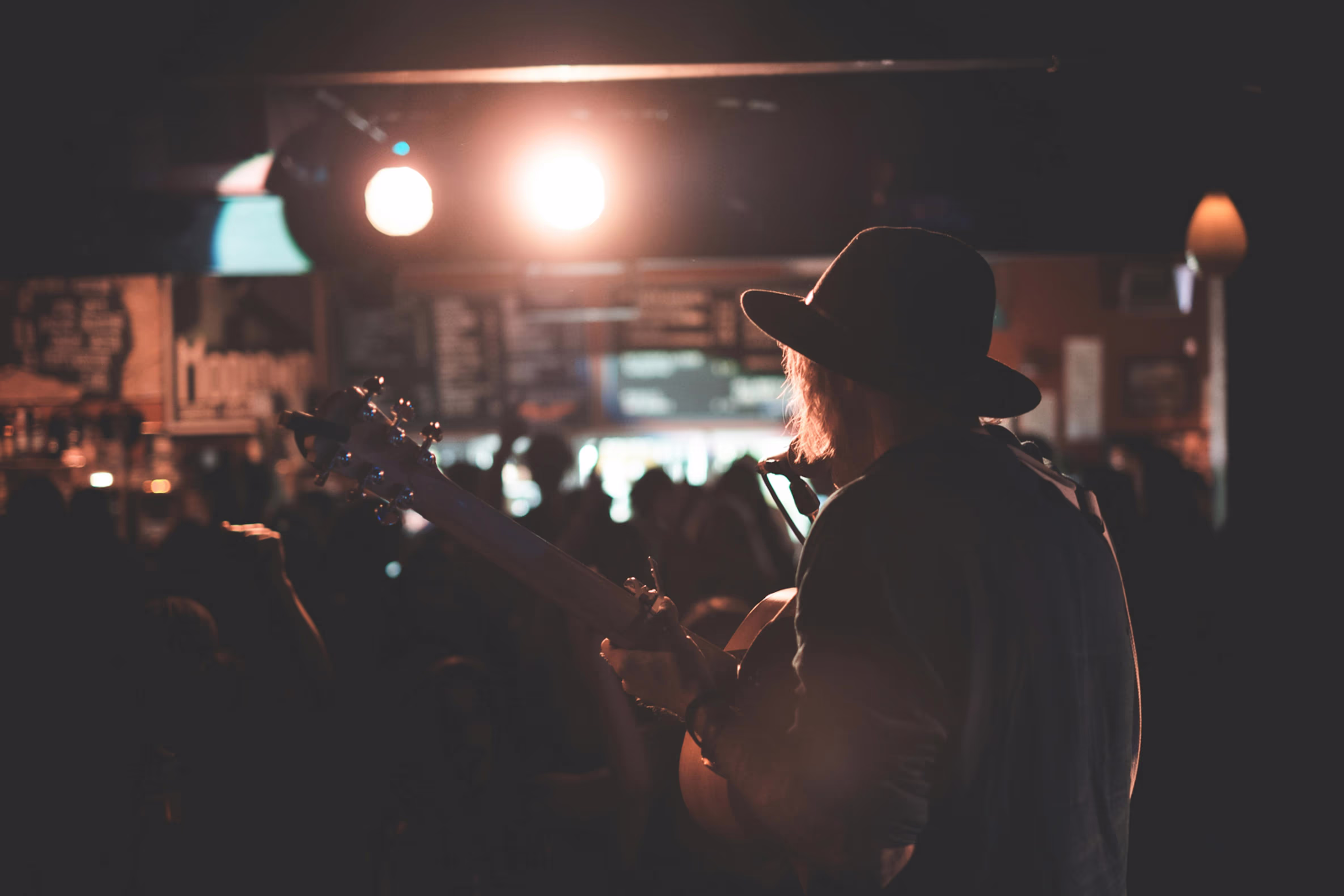Man wearing a hat sings into a microphone while playing a guitar in a dimly lit bar 