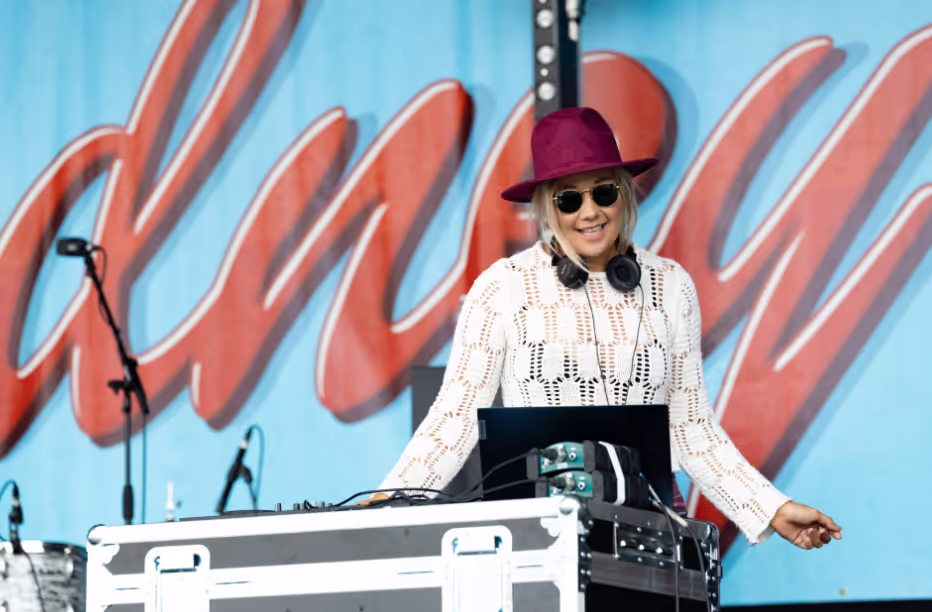 A woman wearing a hat and sunglasses skillfully performing a DJ set at an event, surrounded by vibrant lights.