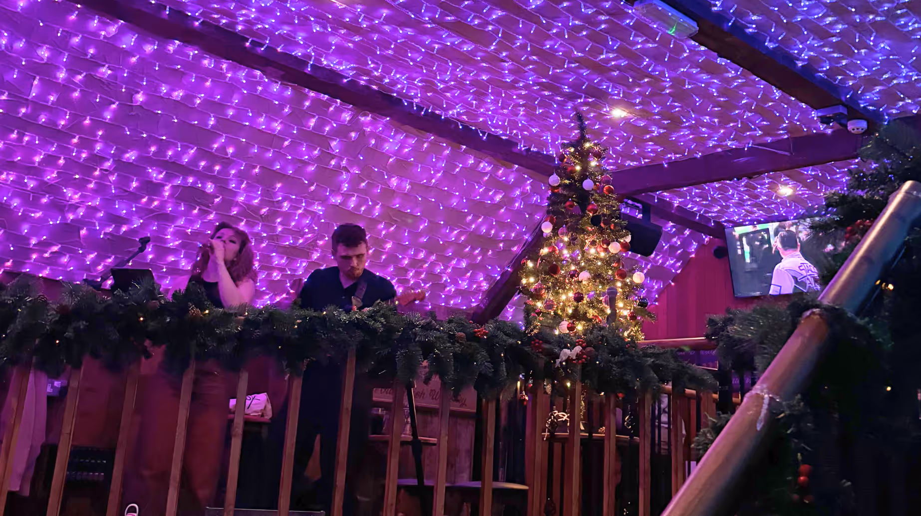 A duo perform in a venue decorated for Christmas, under a ceiling covered in fairy lights