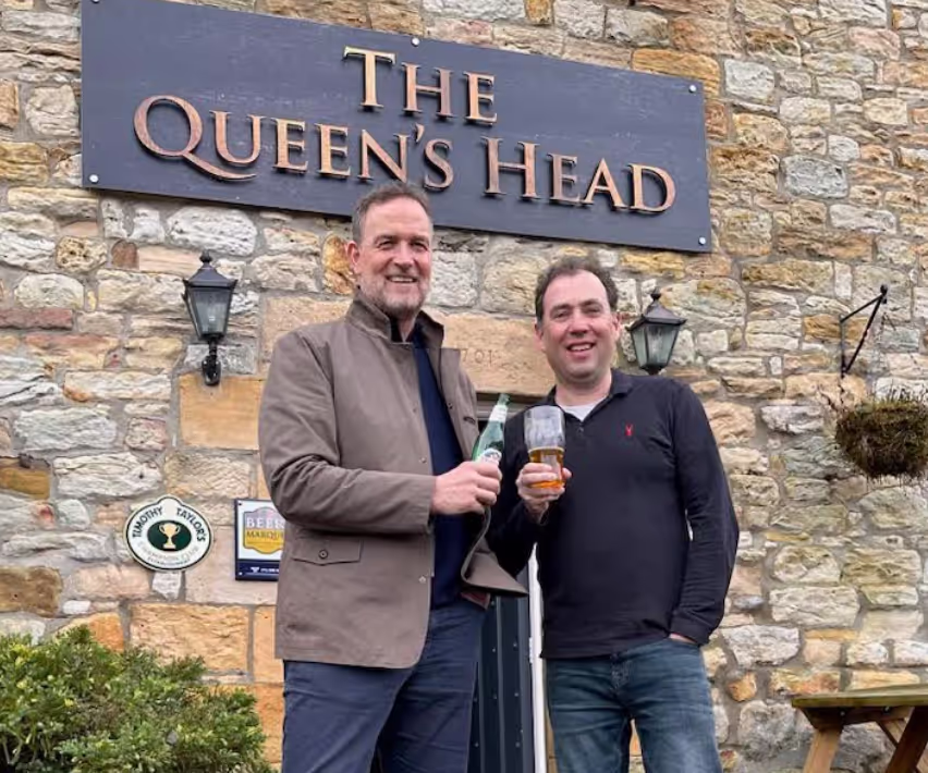 Two men smile into the camera as they stand in front of a pub called The Queens Head, both have drinks in hand 