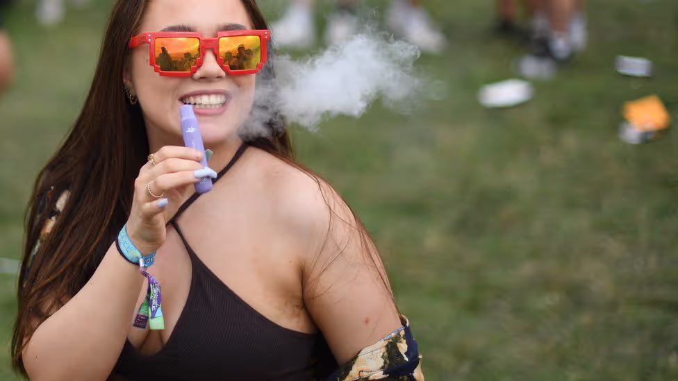 A woman wearing vibrant red sunglasses smiles at a festival as she smokes a vape