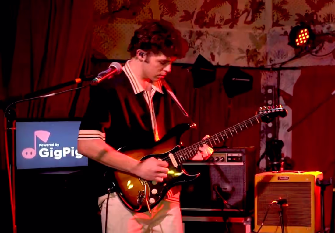 A man plays guitar on stage, with a stack of amps and a GigPig sign behind him