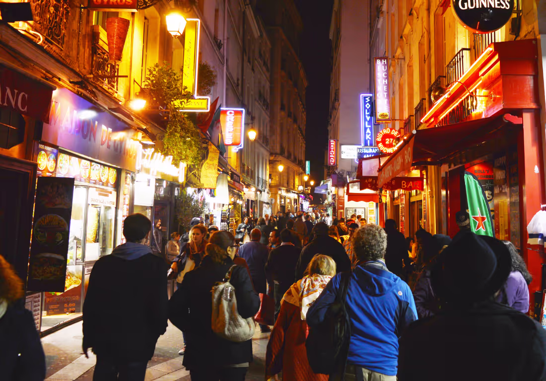 A large crowd of people walk down a brightly lit street at night