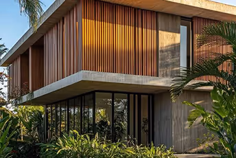 Modern two-story house with large glass windows and wooden slatted panels surrounded by tropical plants.