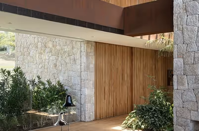 Modern entrance featuring large wooden double doors set between stone walls, surrounded by green plants.