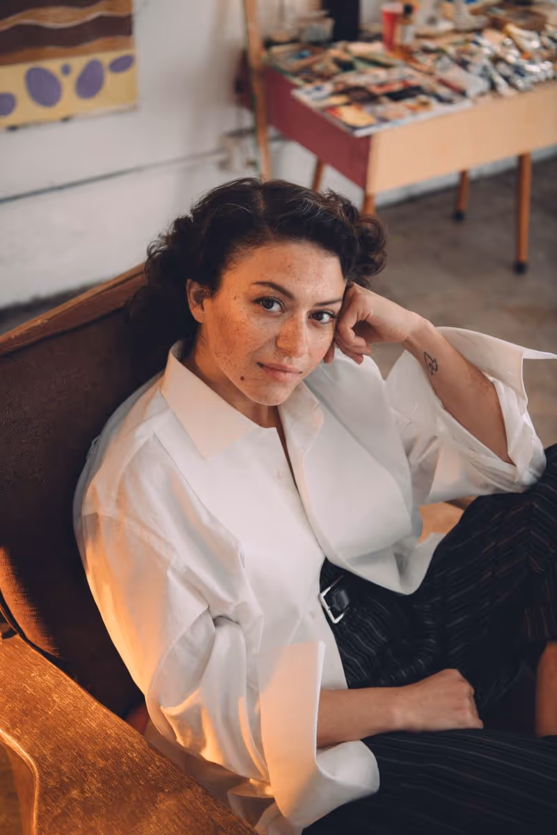 Woman with curly hair and freckles wearing a white shirt sitting on a brown chair and resting her head on her hand.