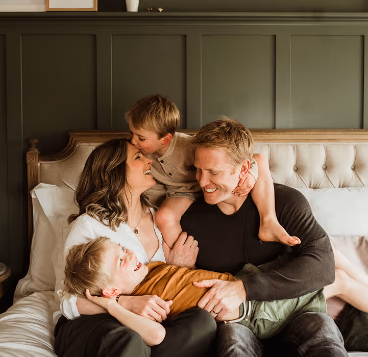 A smiling family of four sitting on a bed, with a mother and father holding two young boys, one kissing the mother’s forehead.