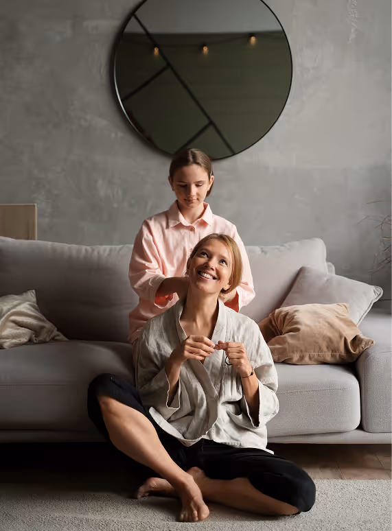 Child standing behind a seated smiling woman, gently touching her hair in a cozy living room.