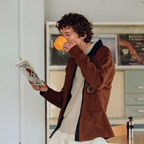 Person with curly hair wearing glasses and a brown jacket drinks from an orange cup while reading a book indoors.