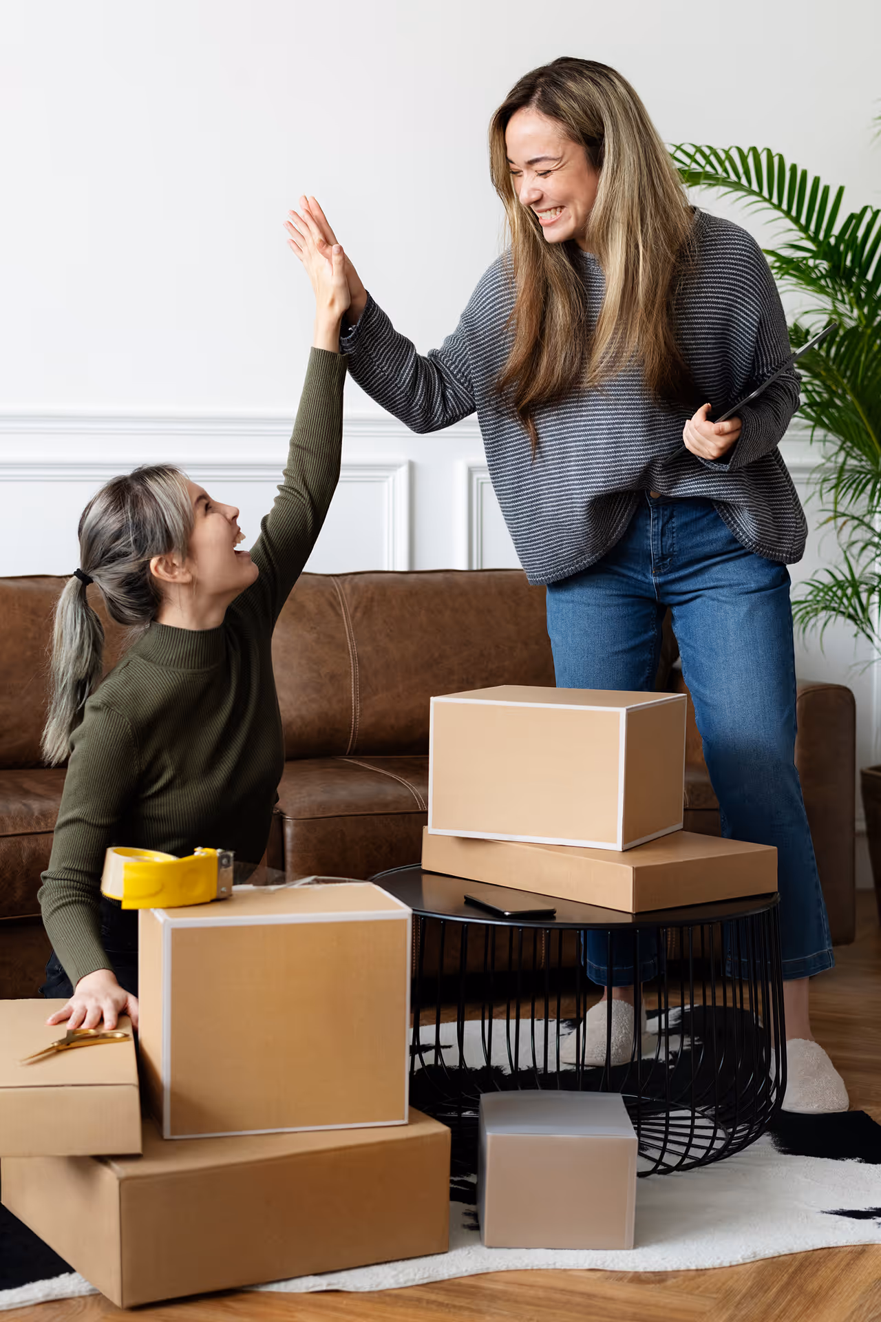 Two women smiling and giving each other a high five while packing cardboard boxes in a living room.