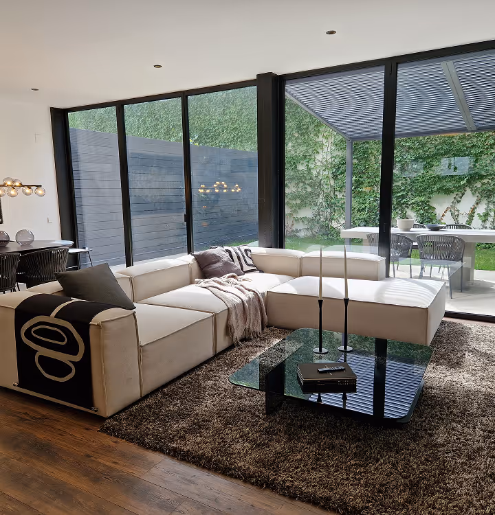 Moderna sala de estar con un gran sofá seccional de color beige, una manta gris oscuro, una mesa de café de vidrio con dos velas y ventanales del piso al techo con vista a un área verde al aire libre.