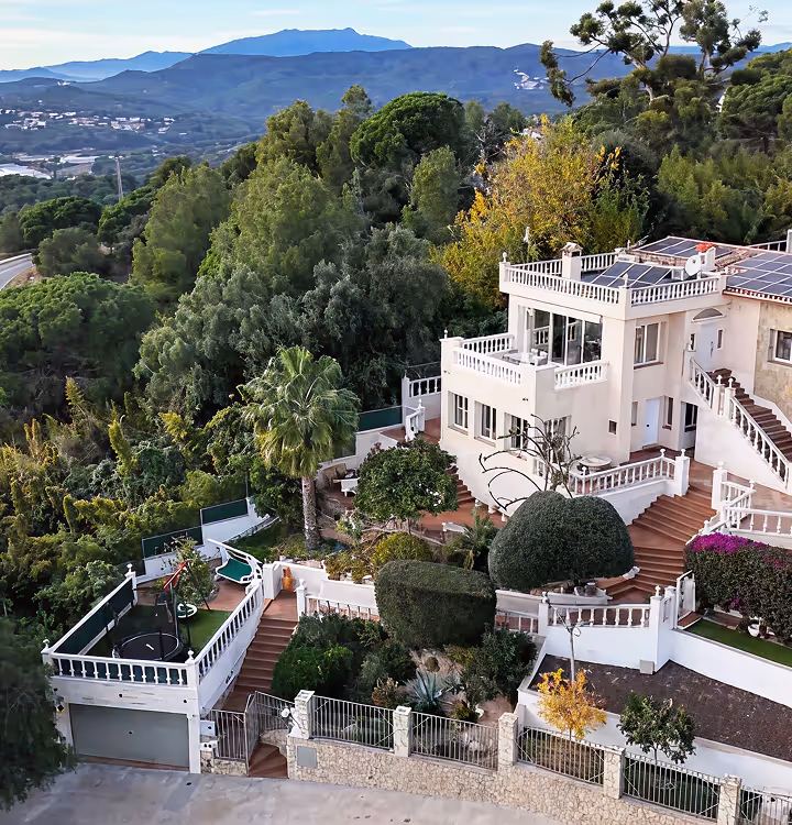 Gran villa blanca con terrazas y paneles solares rodeada de frondosos árboles verdes y vistas a la montaña al fondo.