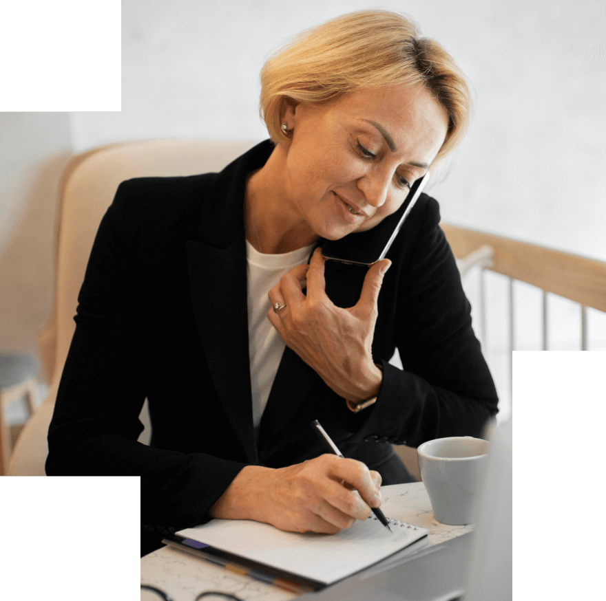 Smiling woman holding a phone between her ear and shoulder while writing in a notebook at a table with a coffee cup.