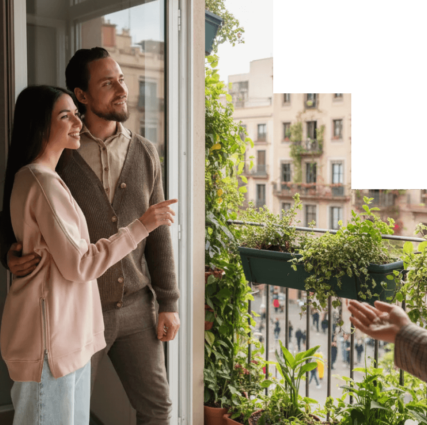 A smiling couple standing by an open window looking out at a balcony garden with plants, while a person’s hand gestures towards the balcony view.