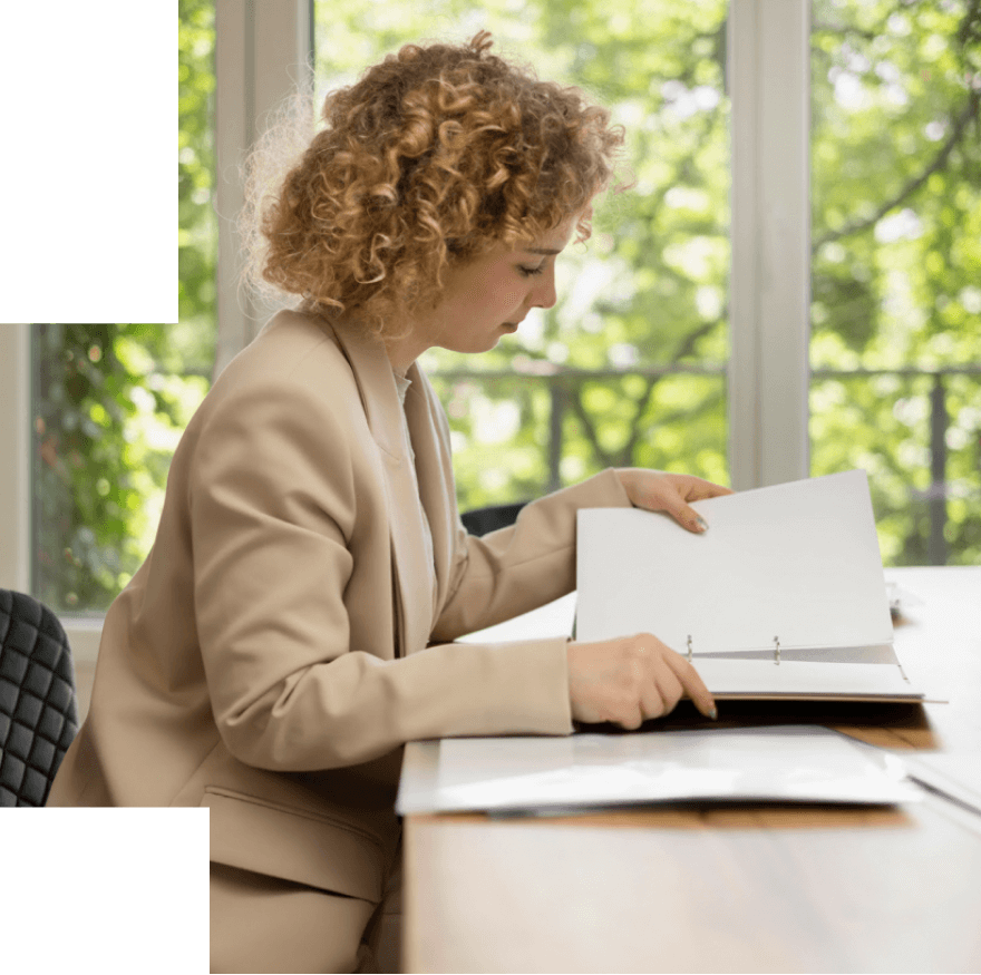 Curly-haired woman in beige blazer sitting at a wooden table, examining pages in a ring binder.