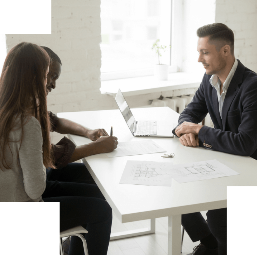 Couple signing documents at a table while a man in a suit watches, with a laptop and floor plans on the table.