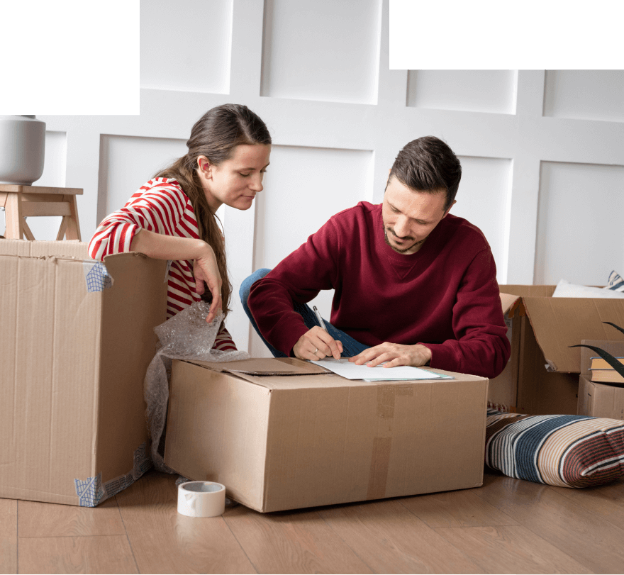 Man writing on a document placed on a cardboard box while a woman in a red and white striped shirt watches, surrounded by moving boxes and packing materials.
