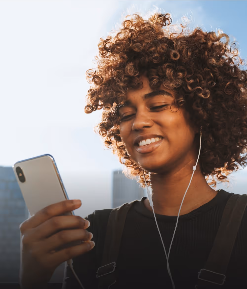Visual for slide: smiling woman with curly hair wearing earphones and looking at her smartphone outdoors.