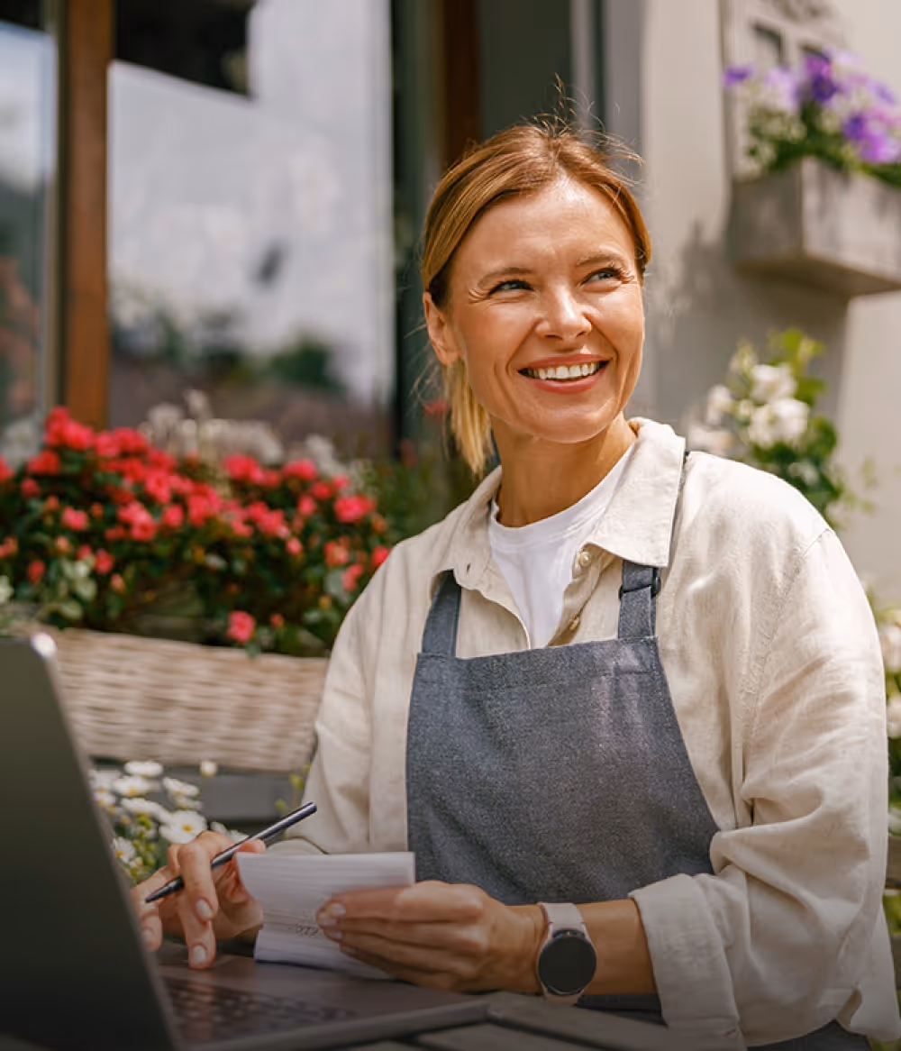 Visual for slide: smiling woman in a gray apron holding a notepad and pen, sitting outdoors with flowers, using a laptop.