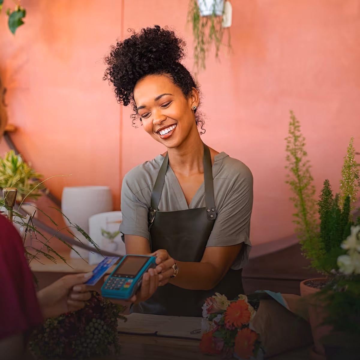 Smiling florist in apron holding a card reader as a customer pays with a credit card at a flower shop counter.