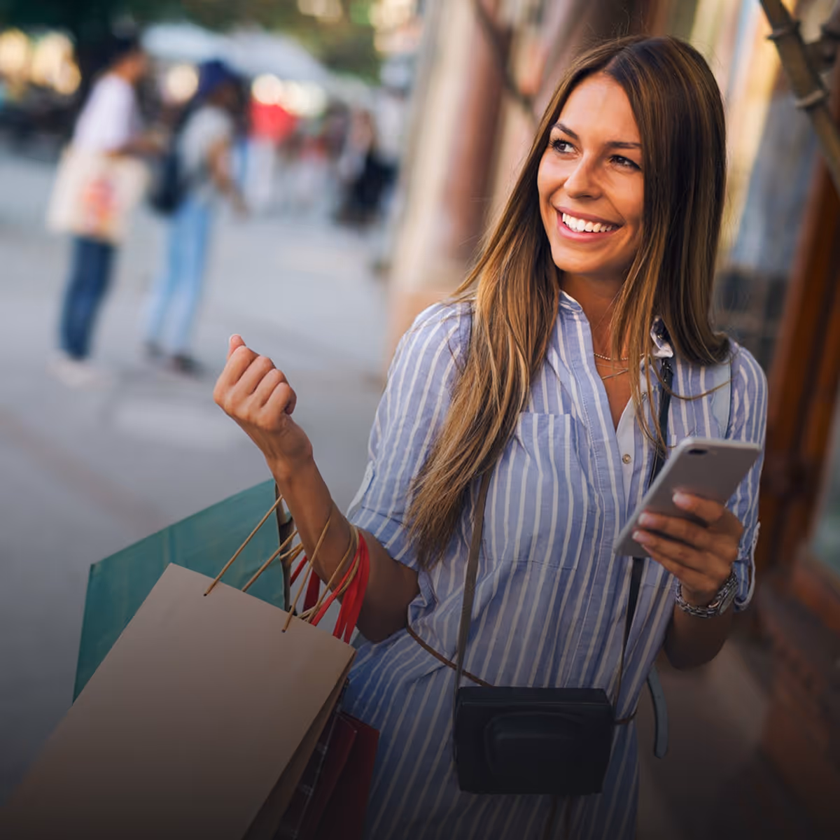 Smiling woman holding shopping bags and using a smartphone outdoors.