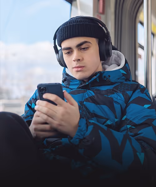 Visual for slide: young man wearing a black beanie and headphones, looking at his phone while sitting indoors by a window.