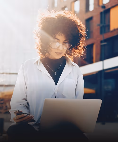 Visual for slide: woman with curly hair and glasses working on a laptop outdoors with sunlight behind her.