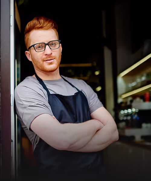 Visual for slide: red-haired man with glasses wearing a black apron and gray shirt, standing with arms crossed in a cafe or restaurant.