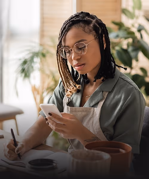 Visual for slide: woman with braided hair and glasses wearing an apron, using a smartphone while writing in a notebook at a table with plant pots.