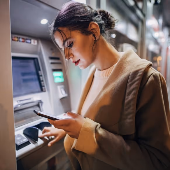 Woman in a beige coat using her smartphone while operating an ATM at night.