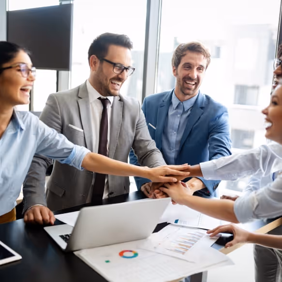 Group of business professionals smiling and stacking hands together over a desk with a laptop and charts.