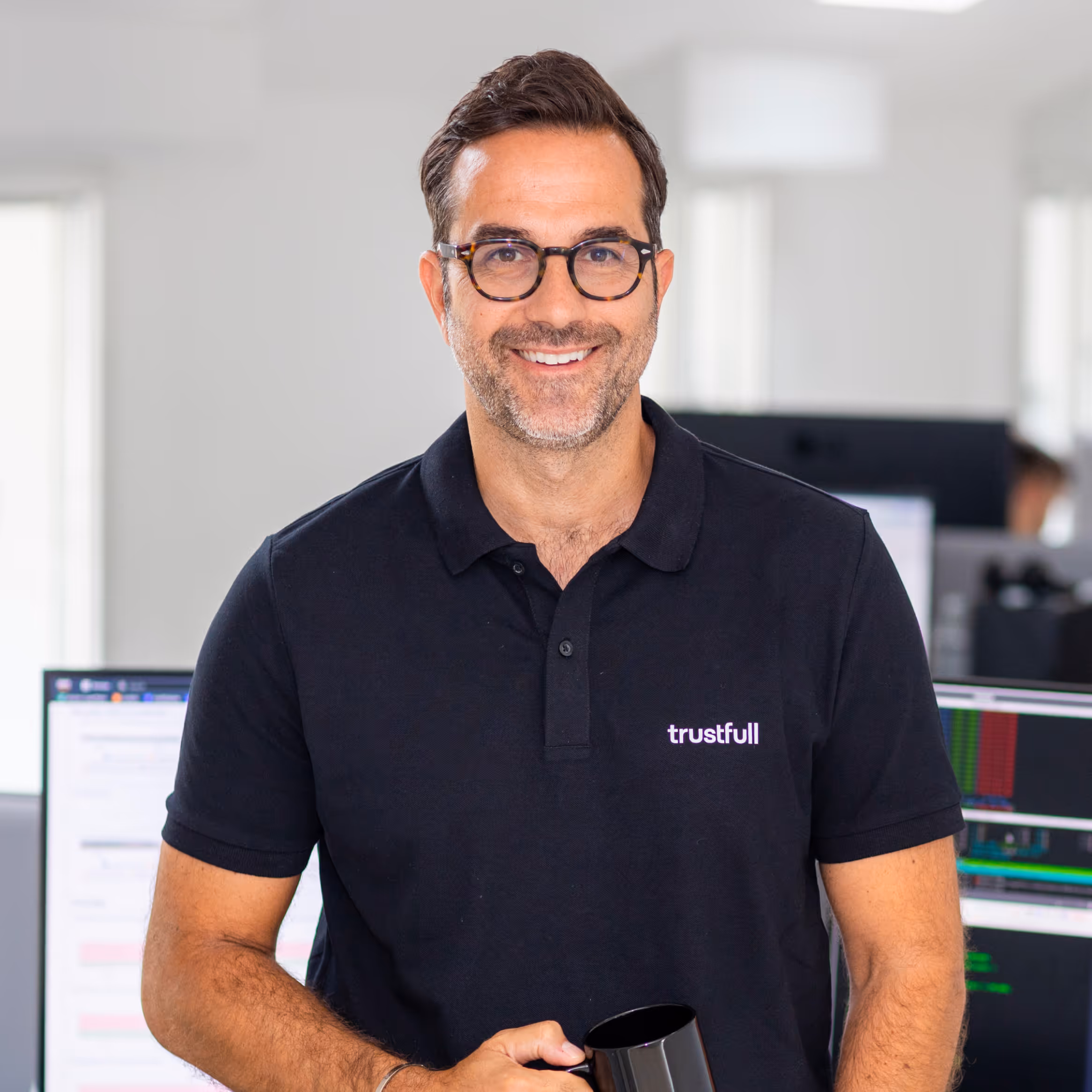 Smiling man with glasses wearing a black polo shirt with 'trustfull' logo, holding a black mug, in an office setting.
