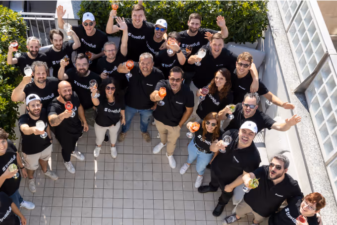 Group of people wearing matching 'trustfull' black shirts raising glasses and smiling outdoors on a tiled patio.