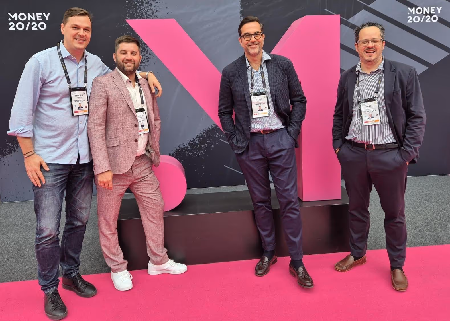 Four men wearing event badges standing and smiling on a pink carpet at the Money 20/20 event with a large pink 20/20 logo in the background.