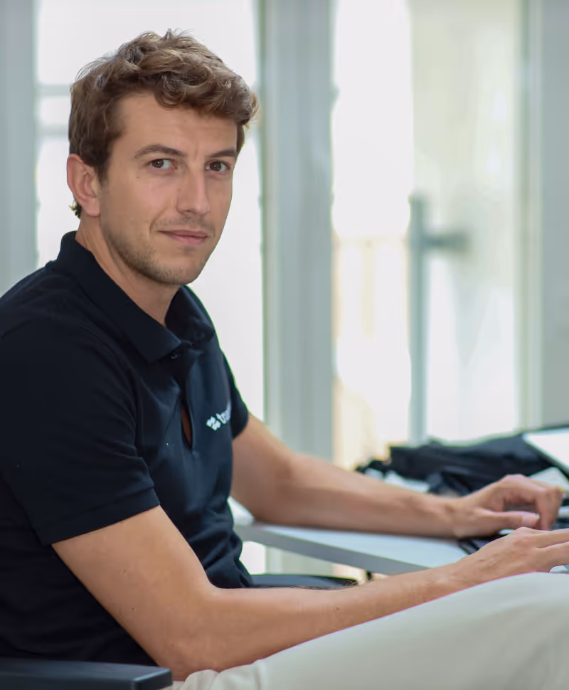Man with curly brown hair seated at a desk, looking towards the camera in a bright room.