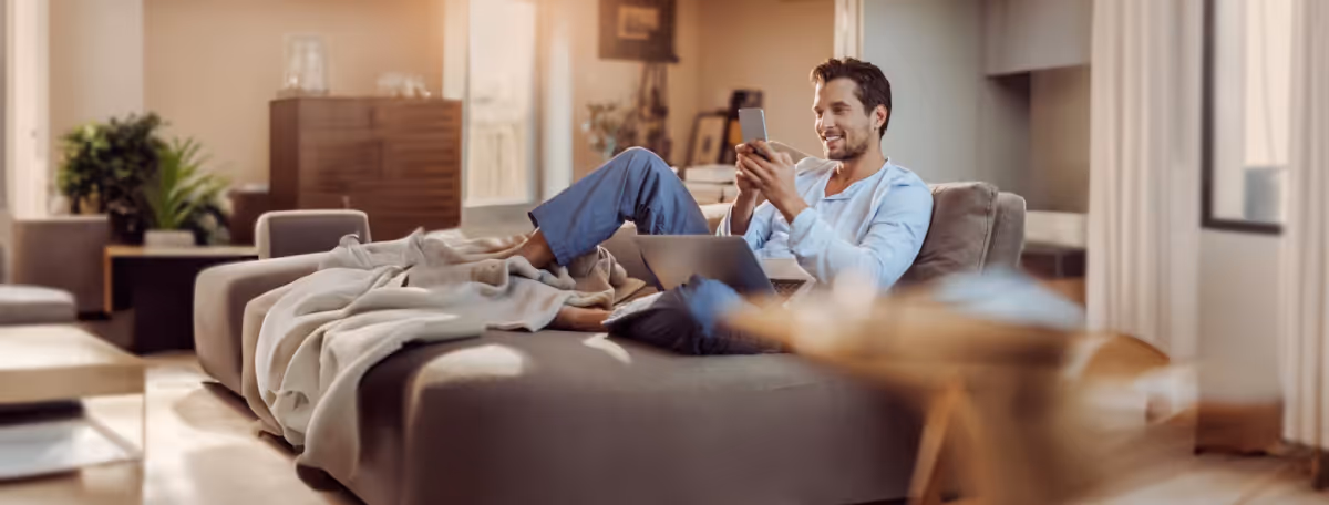 Young man sitting on a sofa in a bright living room, using a smartphone with a laptop on his lap, smiling.