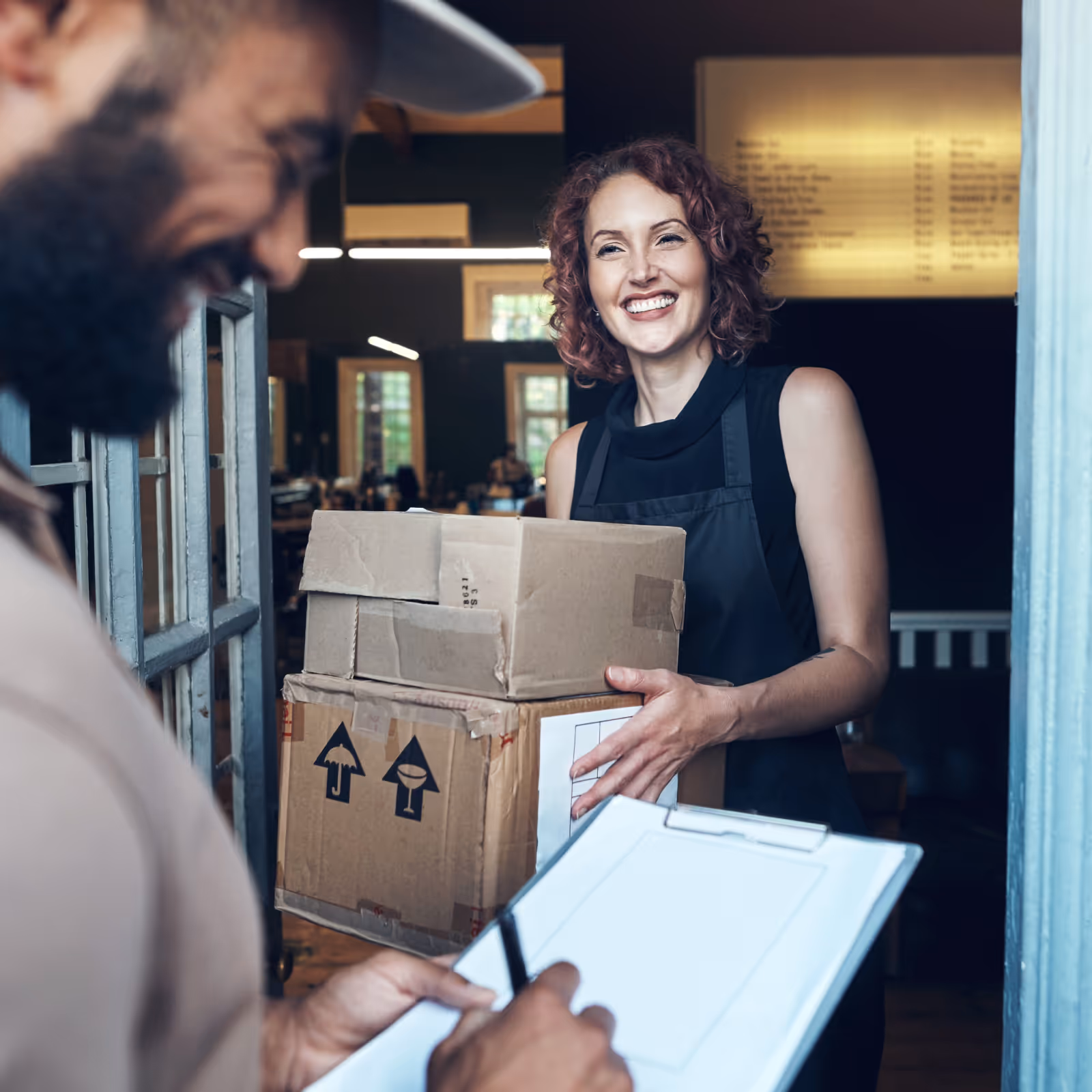 Smiling woman holding delivery boxes at a door while a man signs on a clipboard.