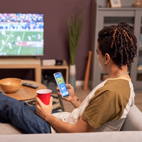 Man sitting on a couch holding a red cup and checking a betting app on his smartphone with a football game on TV in the background.