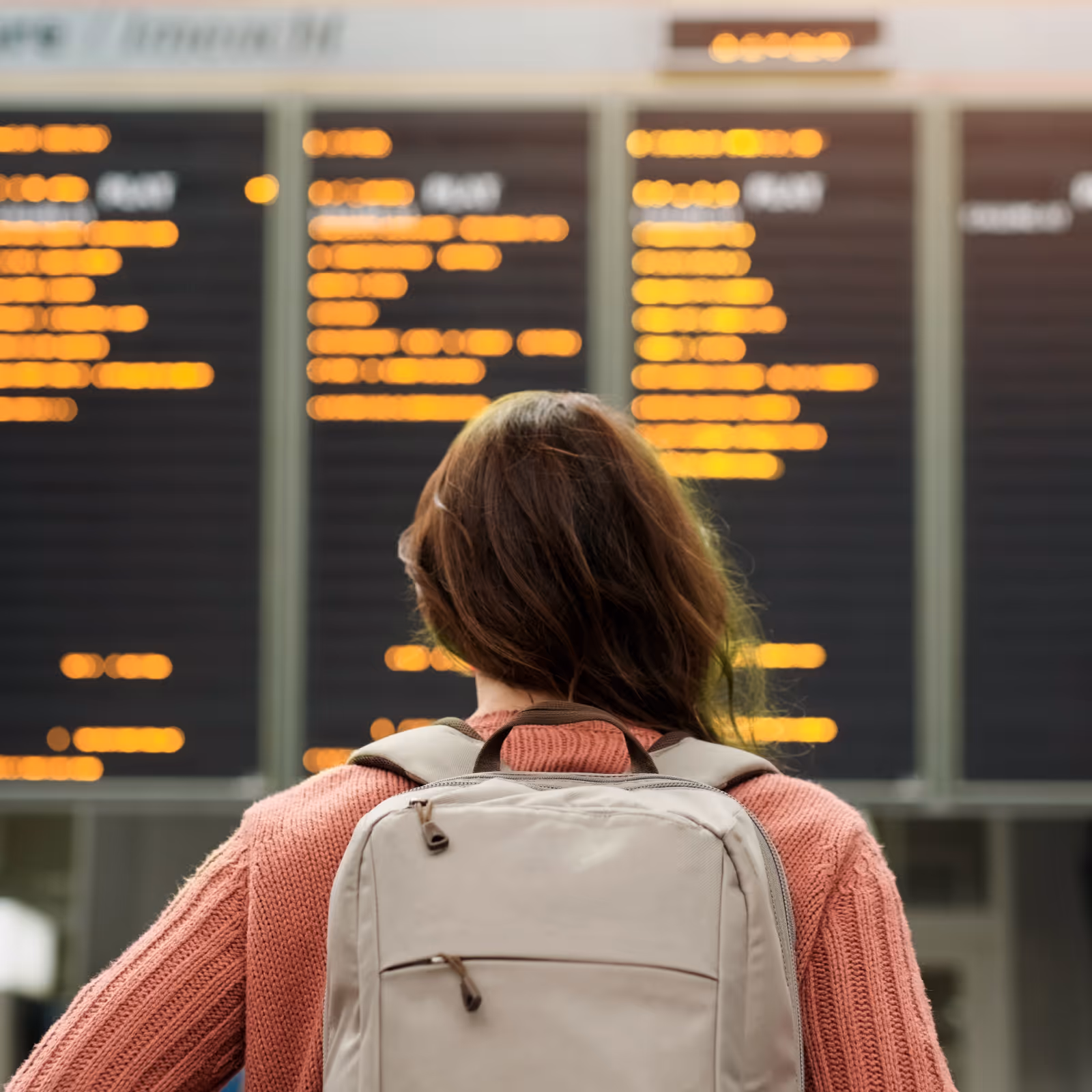 Person with a beige backpack and pink sweater looking at a blurred airport departure board.