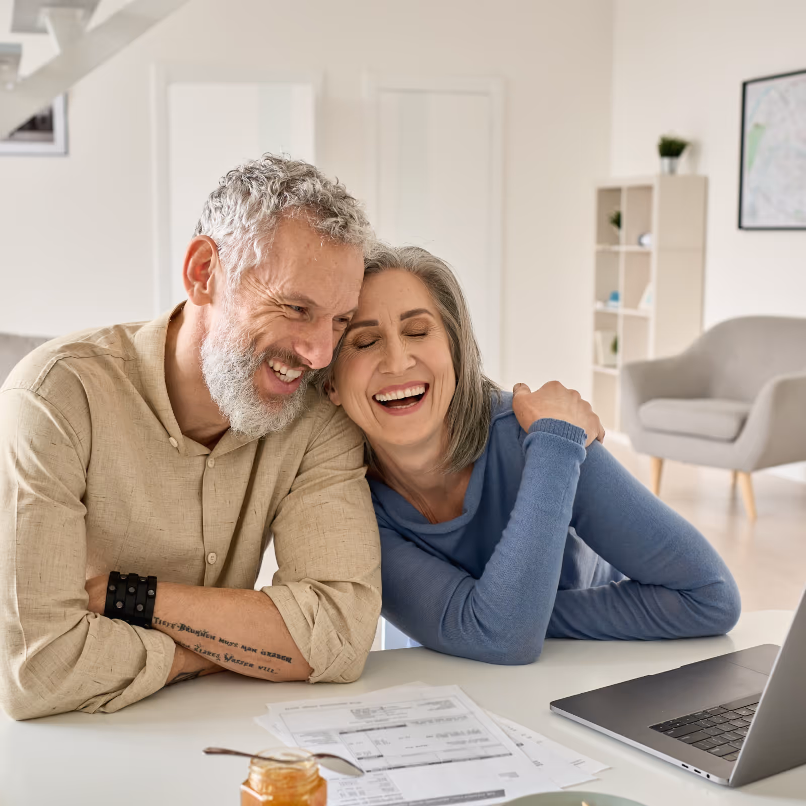 Smiling elderly couple sitting closely together at a table with documents and a laptop in a bright living room.