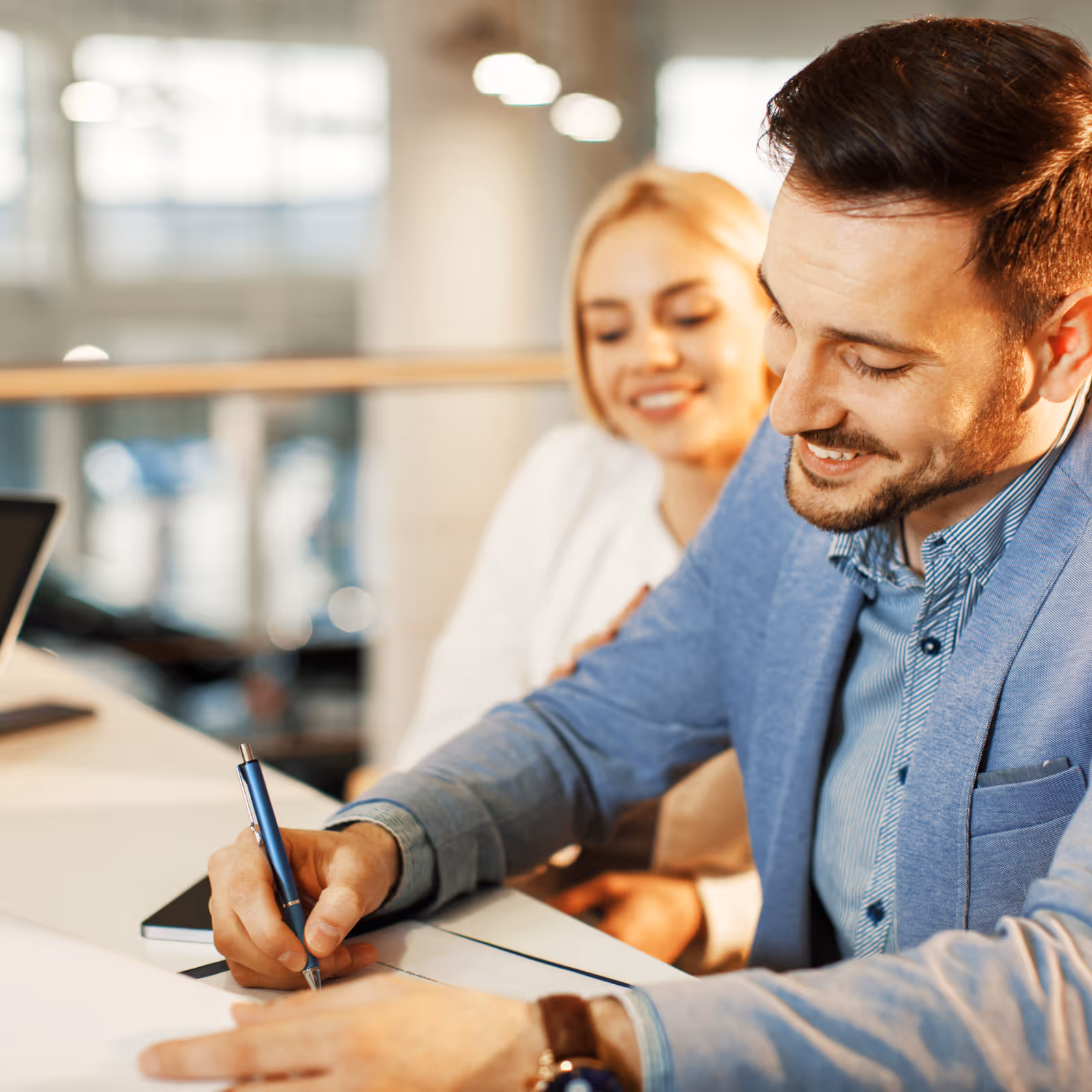 Smiling man in a blue blazer signing a document while a woman watches him.