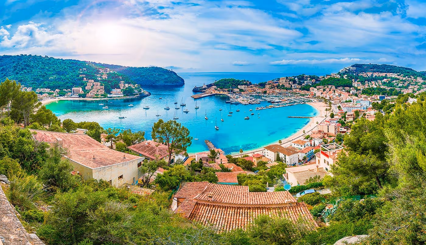 Panoramic view of a coastal town with turquoise bay, anchored sailboats, sandy beaches, and hills covered in greenery.