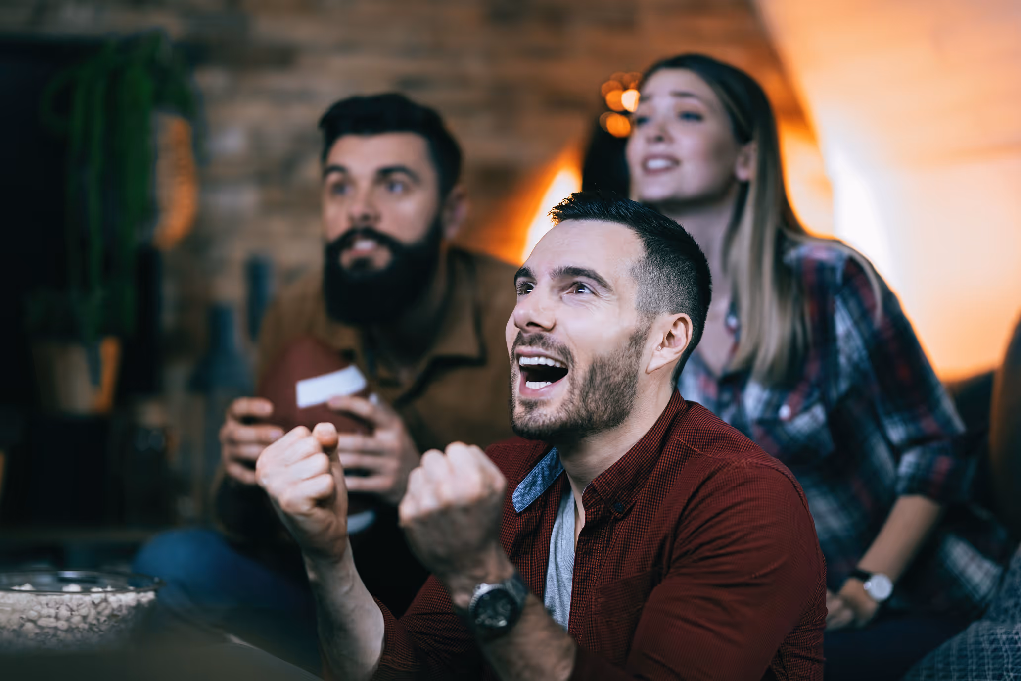 Excited man in red shirt cheering with clenched fists while watching a game with two friends indoors.