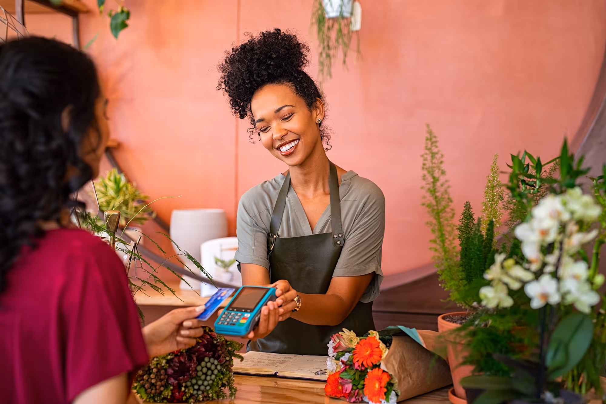 Smiling florist in an apron holding a card machine while customer pays with a credit card in a flower shop.