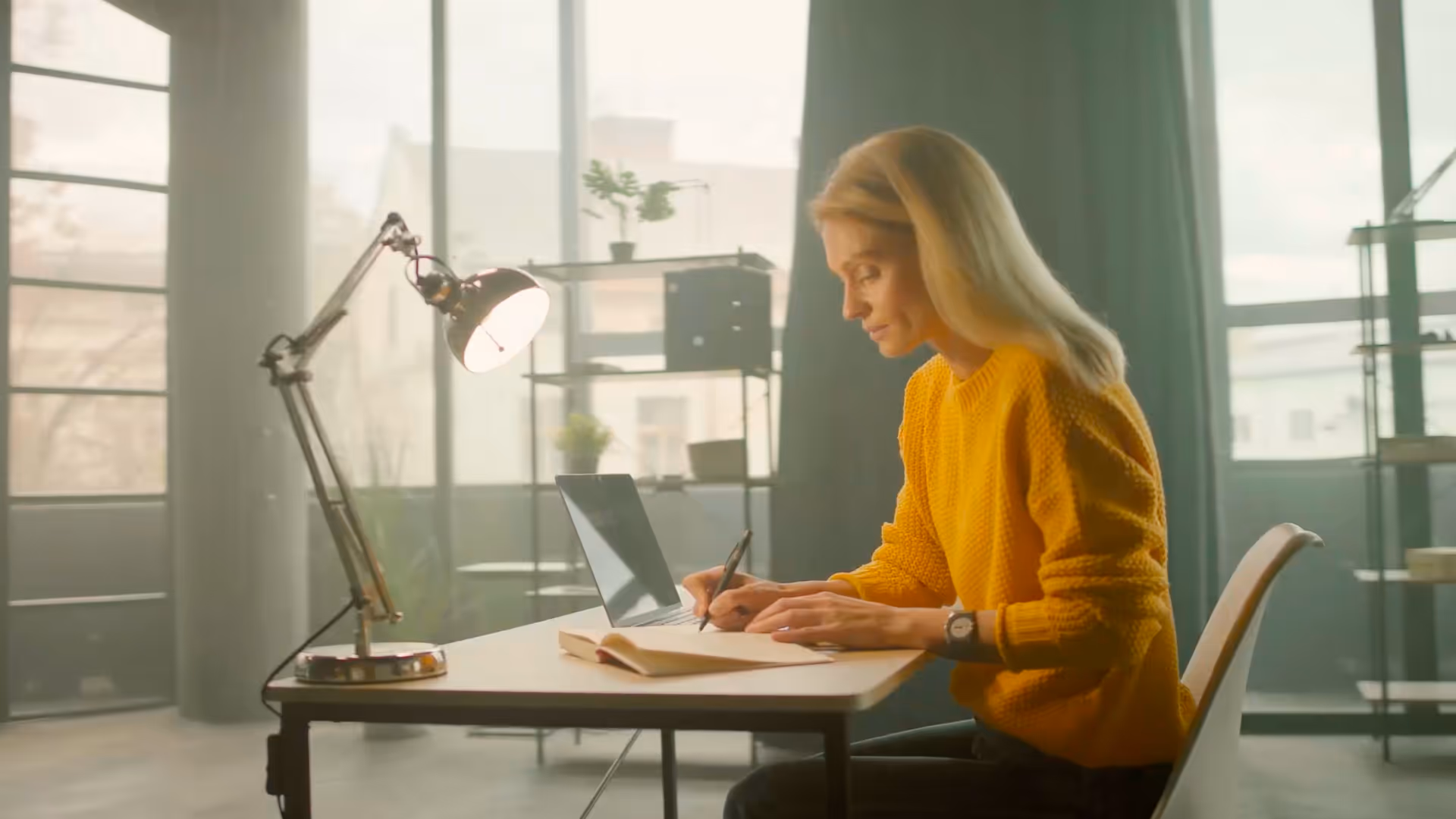 Woman in yellow sweater writing in a notebook at a desk with a laptop and desk lamp in a bright room.