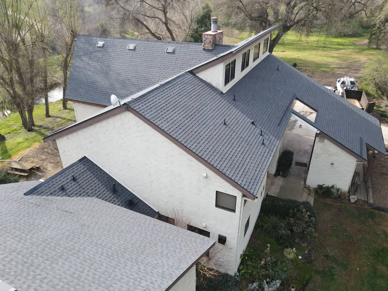 Aerial view of a large residential house with multiple gray shingled roofs and white stucco walls surrounded by greenery.