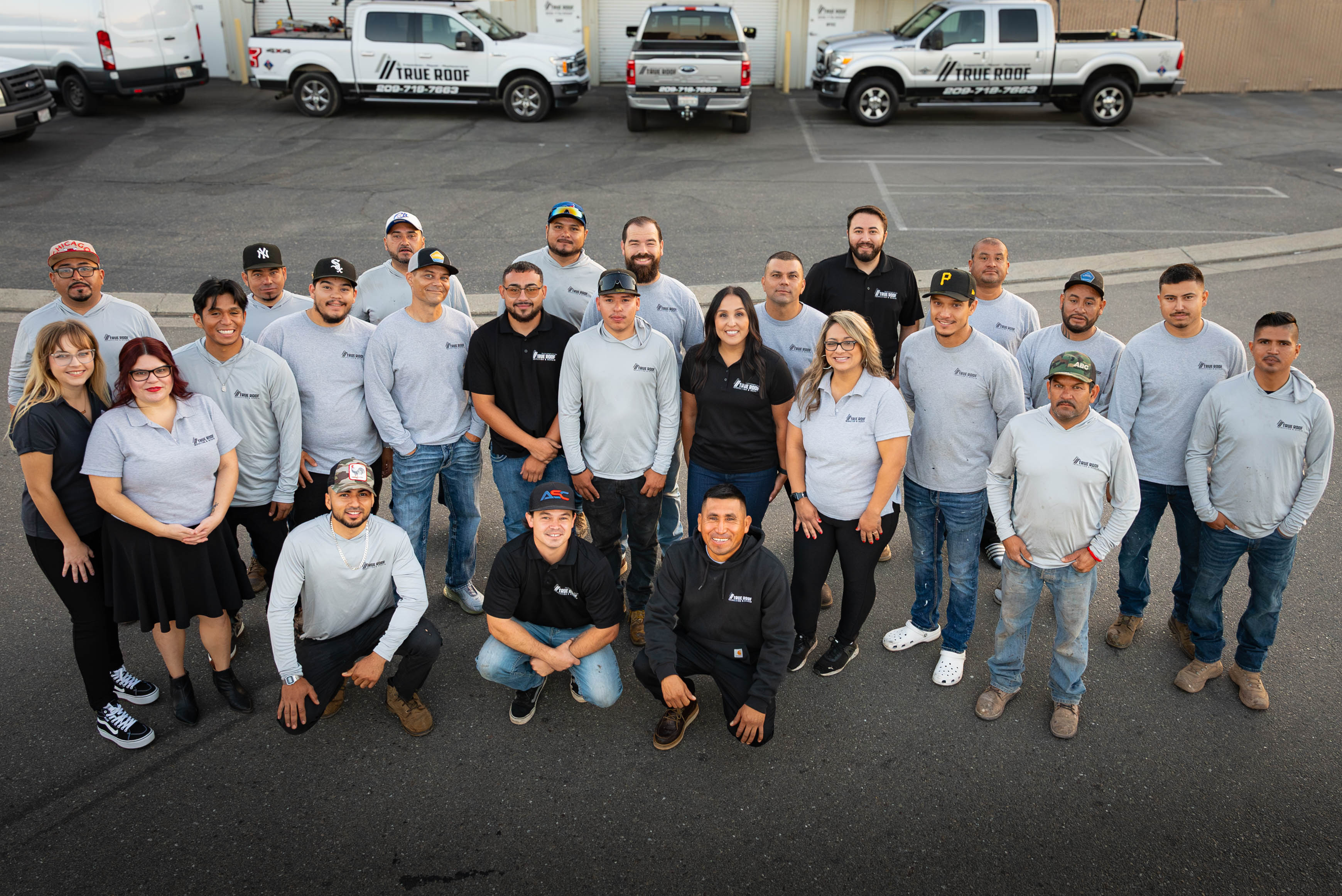 Group photo of 26 True Roof employees standing and kneeling in a parking lot with company trucks in the background.