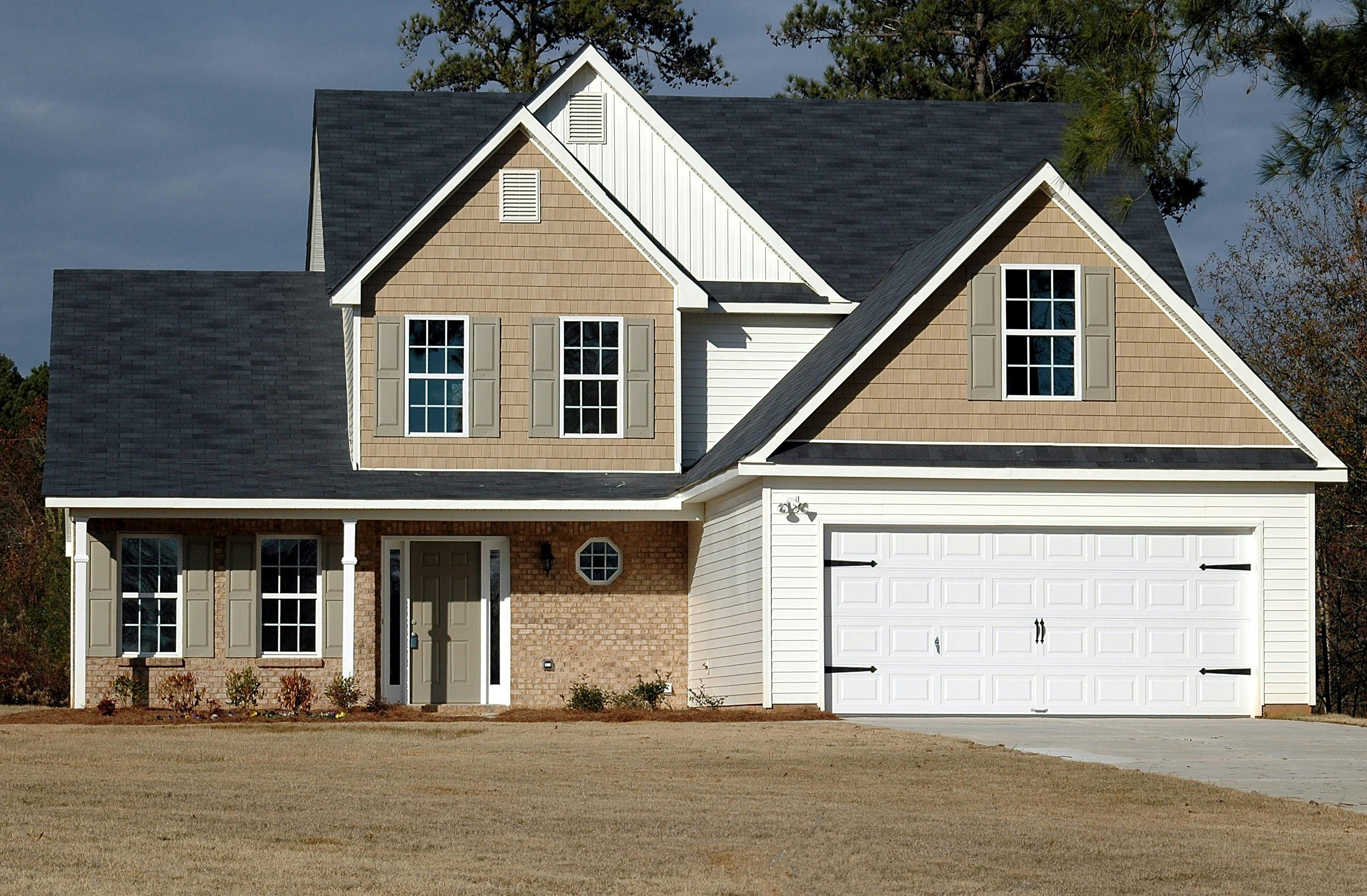 Two-story suburban house with beige siding, white garage door, and a front porch on a dry lawn.