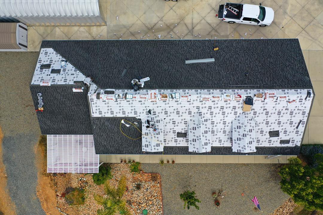 Aerial view of a house roof partially covered with roofing underlayment and workers installing shingles, with a white truck parked on the driveway.